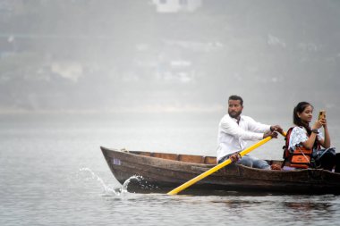 Nanital, Uttarakhand, India - circa 2022 : local man in a colorful traditional cap rowing an elegant row boat with seats for tourists to travel in the foggy lake of Naini jheel in this famous tourist