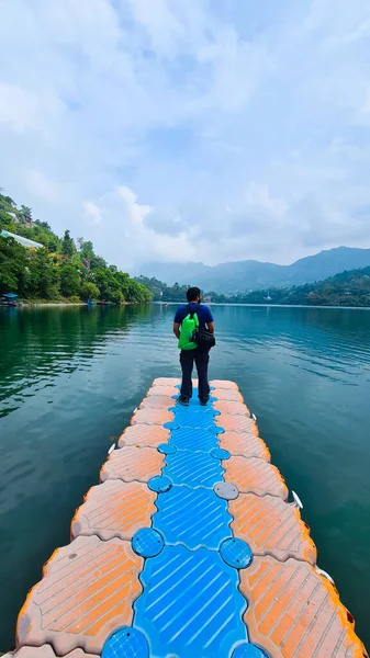 young man standing on plastic pier with the stunning view of mountains fog, clouds and trees over the blue water of naukuchiatal bhimtal lake with a green backpack looking into the distance in India