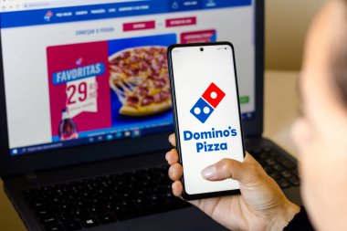 18 Nisan 2022, Brezilya. Bu resimde, Domino 's Pizza logosunun ekranda sergilendiği bir akıllı telefonu olan bir kadın arka planda Domino' s Pizza web sitesi var.