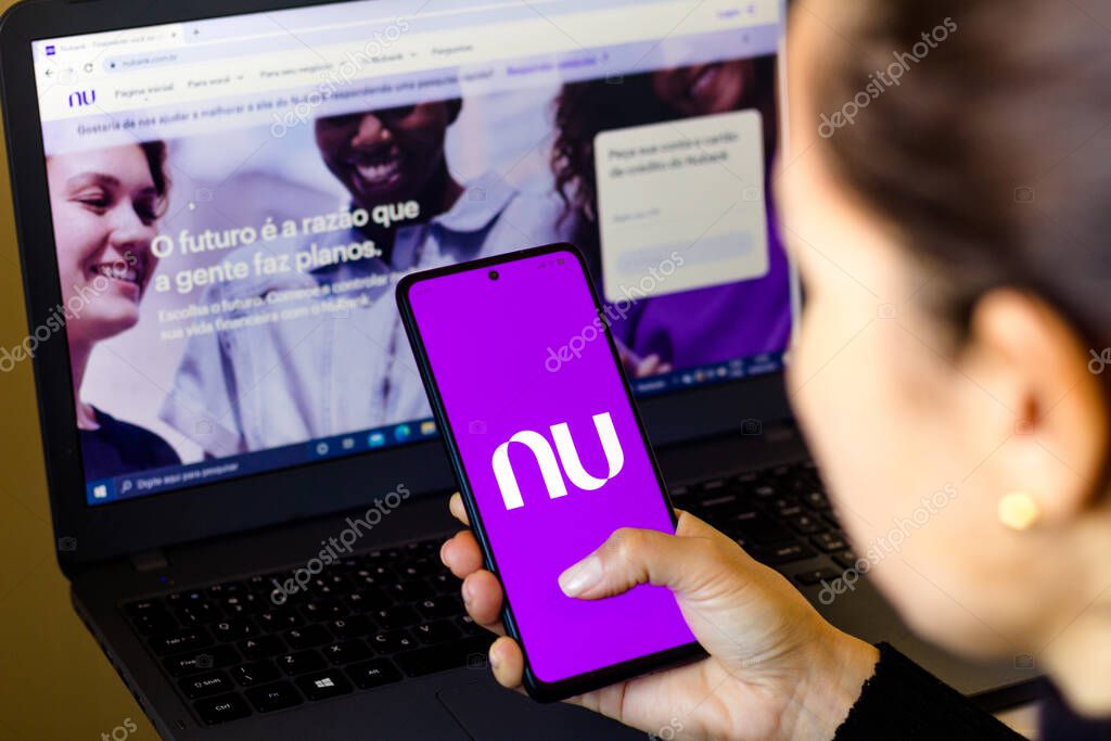 April 18, 2022, Brazil. In this photo illustration, a woman holds a smartphone with the Nubank logo displayed on the screen with the Nubank website displayed in the background