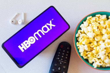 19 Nisan 2022, Brezilya. Bu resimde, akıllı telefondaki HBO Max logosu bir kase patlamış mısır, kulaklık ve uzaktan kumandayla birlikte görülüyor.