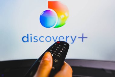 May 10, 2022, Brazil. In this photo illustration a close-up of a hand holding a TV remote control seen in front of the Discovery Plus logo