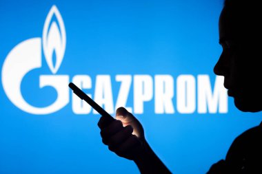 June 29, 2022, Brazil. In this photo illustration, the Gazprom logo is seen in the background of a silhouetted woman holding a mobile phone