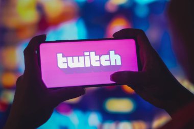 July 6, 2022, Brazil. In this photo illustration, a silhouetted woman holds a smartphone with the Twitch logo displayed on the screen