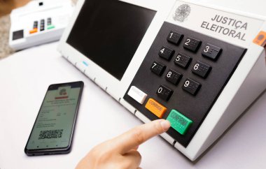2 Ekim 2022, Brezilya. Bu fotoğraf illüstrasyonunda, Brezilya 'da seçimlerde kullanılan elektronik oy sandığı (oy sandığı) içinde oy kullanma simülasyonunu bir kişi yapar. Masanın yanında bir akıllı telefon var.