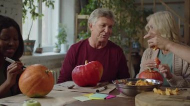 İnsanlar Cadılar Bayramı için hazırlanıyor. Masada otururlar, sohbet ederler, balkabağı yerler ve lamba keserler.