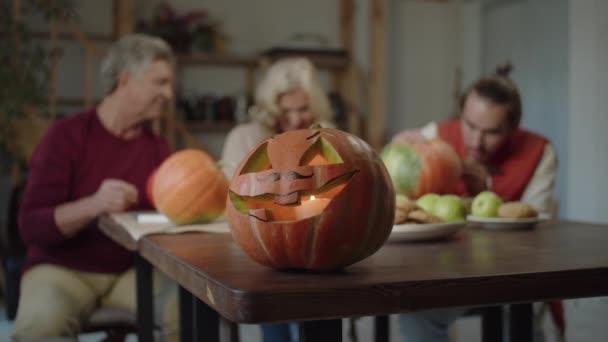 Am Tischrand steht eine Kürbislaterne mit einer Kerze im Mund. Menschen im Hintergrund bereiten sich eifrig auf Halloween vor — Stockvideo