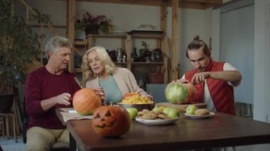  Genç bir adam ve büyükannesi ve büyükbabası masada oturup Cadılar Bayramı için balkabağı oyuyorlar. Güzel iletişim kuruyorlar.