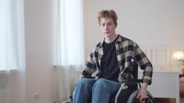 Ένας νεαρός κάθεται σε αναπηρικό καροτσάκι και κοιτάει σοβαρά την κάμερα. — Αρχείο Βίντεο