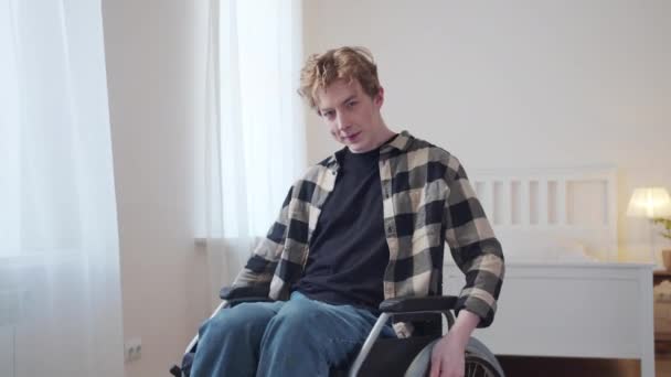 Een jonge gehandicapte man rijdt in zijn rolstoel door de kamer en kijkt naar de camera — Stockvideo