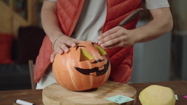 Un joven con chaleco rojo talla cejas de una calabaza de halloween — Vídeos de Stock