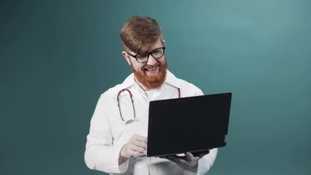 Un joven médico barbudo rojo está hablando con su amigo en Internet y sonriendo — Vídeos de Stock
