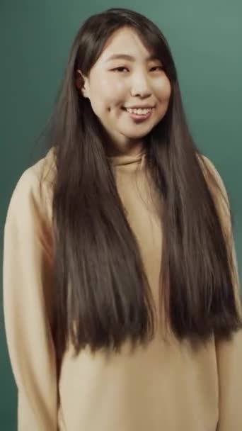 Ελκυστική χαρούμενη Κινέζα με μακριά μαλλιά ποζάροντας για την κάμερα, χαμογελώντας και κουνώντας τα χέρια — Αρχείο Βίντεο