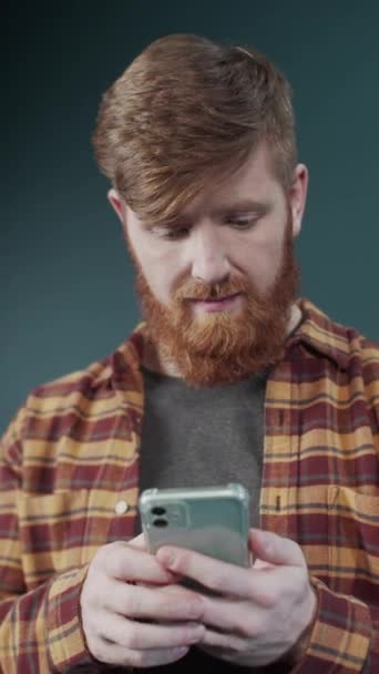 누런 셔츠를 입고 녹색 배경에 서서 새로운 스마트폰으로 비디오를 보고 있는 남자 — 비디오