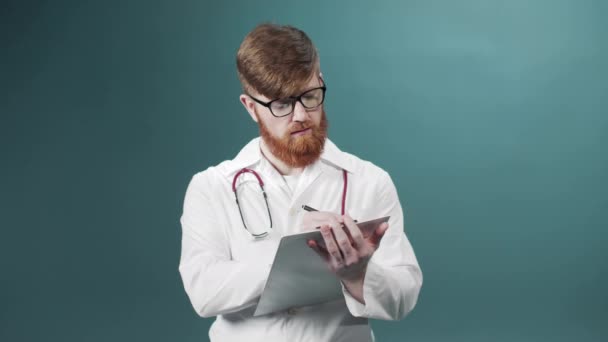 Um jovem médico de vestido branco está escrevendo algo em um tablet guardando em suas mãos — Vídeo de Stock