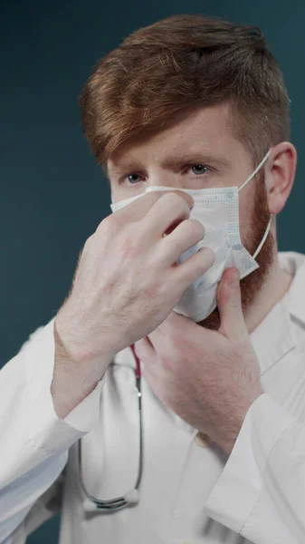 Hombre con barba con cara seria en bata de laboratorio pone máscara protectora en aislado sobre fondo de laboratorio oscuro —  Fotos de Stock