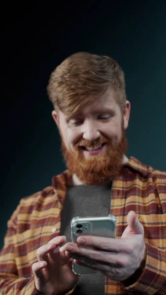 Рыжий парень в современной клетчатой рубашке смотрит в экран нового популярного мобильного телефона — стоковое фото