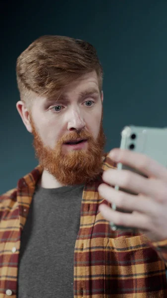 Entusiástico jovem ruiva homem fazer uma chamada de vídeo por câmera de telefone perto de uma parede verde — Fotografia de Stock