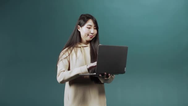 Una hermosa chica asiática en sudadera con capucha beige está utilizando su computadora portátil para charlar con amigos, sonriendo y mostrando un pulgar hacia arriba — Vídeos de Stock