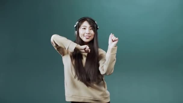 Милая очаровательная китаянка в бежевой толстовке с наушниками, танцующими на цветном фоне — стоковое видео