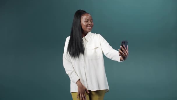 Uma menina africana bonita em uma blusa branca está tirando uma selfie, escolhendo fotos e sorrindo — Vídeo de Stock
