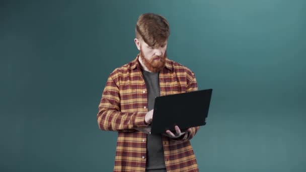 Młody facet z rudymi włosami i brodą ujawnia poważne nastawienie trzymając laptopa — Wideo stockowe