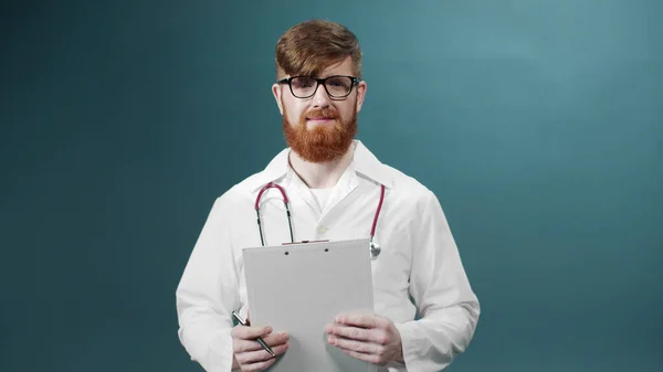 Un giovane dottore in occhiali posa in camice bianco con penna e referto medico in mano — Foto Stock