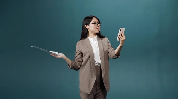 Una guapa jovencita asiática en traje de oficina y gafas con documentos en sus manos hablando en la videollamada con su amiga —  Fotos de Stock