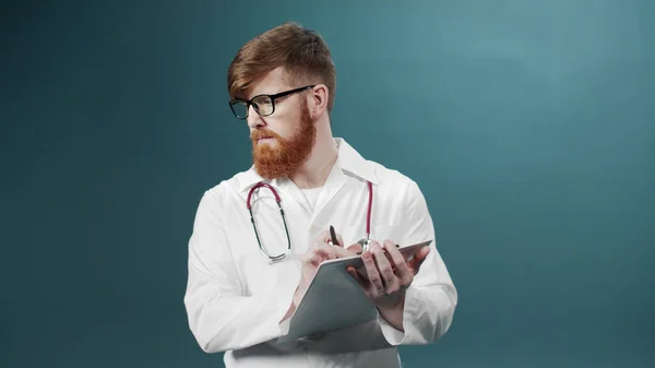 A young doctor in a white gown is writing something down on a tablet keeping in his hands — Stock Photo, Image
