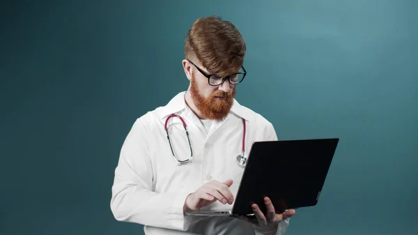 Een volwassen arts in een medisch uniform gebruikt zijn laptop om door het internet te zoeken. — Stockfoto
