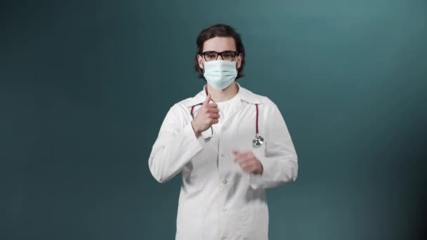 Белый мужчина с черными волосами в медицинском халате и защитной маске стоит с большим пальцем вверх и смотрит в камеру — стоковое видео