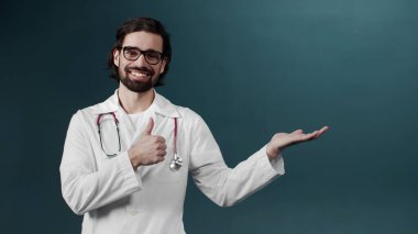 Siyah saçlı, siyah elbiseli beyaz bir adam poz veriyor, başparmağıyla kameraya bakıyor.