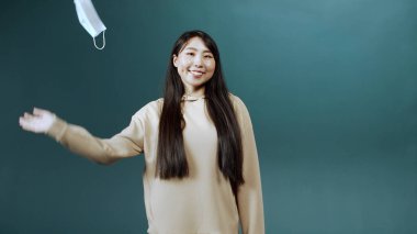 Uzun saçlı Asyalı bir kız koruyucu yüz maskesini çıkarıyor ve gülümsüyor.