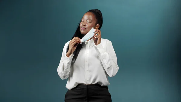 Uzun saçlı, koruyucu yüz maskesi takan, gülümseyen ve dans eden koyu tenli bir kız.