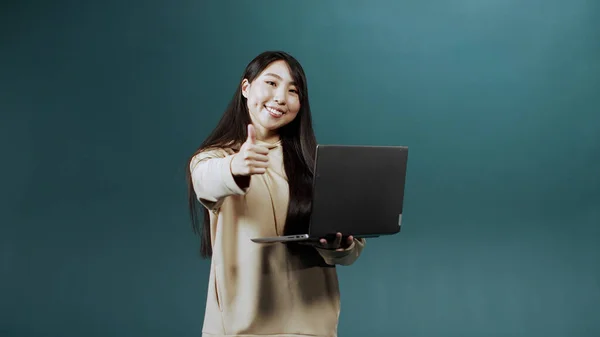 Una hermosa chica asiática en sudadera con capucha beige está utilizando su computadora portátil para charlar con amigos, sonriendo y mostrando un pulgar hacia arriba —  Fotos de Stock
