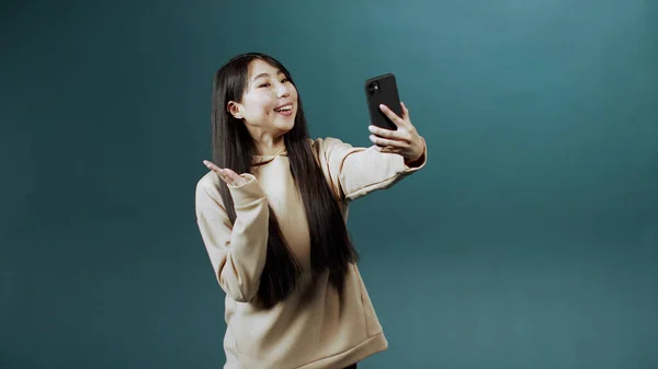Chica china en sudadera con capucha beige mira en la pantalla del nuevo teléfono inteligente, hace selfie, agita la mano —  Fotos de Stock
