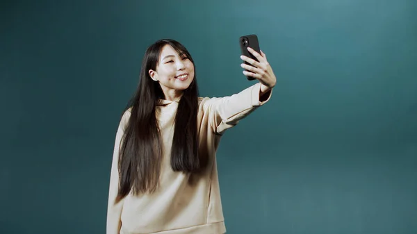 Chica bonita con capucha beige mira en la pantalla del nuevo teléfono móvil, hace selfie, muestra su lengua —  Fotos de Stock