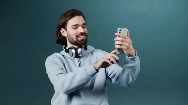 Gri kapüşonlu, kulaklıklı çekici, neşeli adam yeni yeşil akıllı telefonuyla selfie çekiyor. — Stok fotoğraf