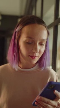 Genç bir kız öğrenci pencerenin yanında duruyor, uzaklara bakıyor ve sonra internette gülümseyen yazılar okuyor. Yüksek kaliteli FullHD görüntüler