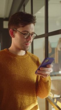 Yakışıklı genç bir öğrenci pencerenin yanında duruyor, elinde bir telefon tutuyor ve düşünceli bir şekilde bir şeyler okuyor. Yüksek kaliteli FullHD görüntüler