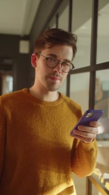 Yakışıklı genç bir öğrenci pencerenin yanında duruyor, elinde bir telefon tutuyor ve kameraya bakıyor. Yüksek kaliteli FullHD görüntüler