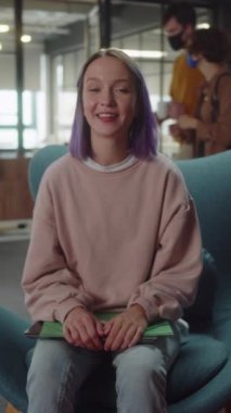 Ön planda genç bir kadın üniversitede yumuşak bir sandalyede oturuyor ve yavaş çekimde kameraya bakıyor. Yüksek kaliteli FullHD görüntüler