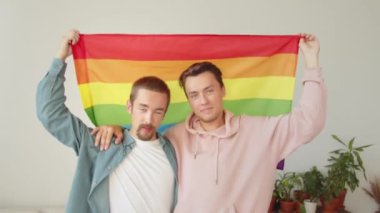 Günlük kıyafetler giymiş iki çekici adam ayakta duruyor ve Igbt bayrağı gösteriyorlar, gülümsüyorlar. Yüksek kaliteli FullHD görüntüler