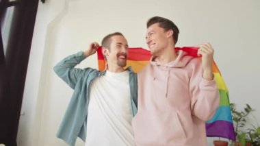Günlük giysiler içinde iki yakışıklı genç ayakta, bir lgbt bayrağını örterek gülümsüyor. Yüksek kaliteli FullHD görüntüler