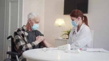 Beyaz önlüklü ve maskeli bir kadın yaşlı bir adama iğne yapıyor. Her şeyin yolunda olduğunu söylüyor. Hastanın baş parmağı görünüyor. Yüksek kalite 4k görüntü