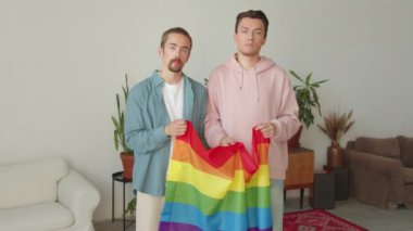 Yetişkin yakışıklı erkekler oturma odasının ortasında durup birbirlerine bakıyorlar ve Igbt bayrağını açıyorlar. Yüksek kaliteli FullHD görüntüler
