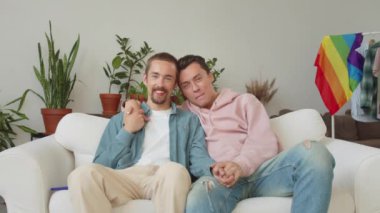 Lgbt genç yakışıklı adamlar kanepede birlikte oturuyorlar, el ele tutuşuyorlar ve kameraya bakıyorlar. Yüksek kalite 4k görüntü