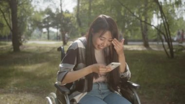 Uzun siyah saçlı, engelli bir Asyalı genç kız halka açık bir parkta tekerlekli sandalyede oturuyor, telefonunda komik bir şeyler okuyor ve gülümsüyor, ağır çekimde. Yüksek kaliteli FullHD görüntüler