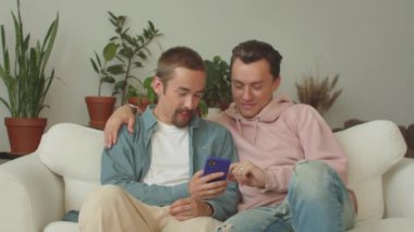 Lgbt adamları oturma odasındaki kanepede oturuyorlar ve internette telefonla bir şeyler izliyorlar, sonra da gülümsüyorlar. Yüksek kalite 4k görüntü