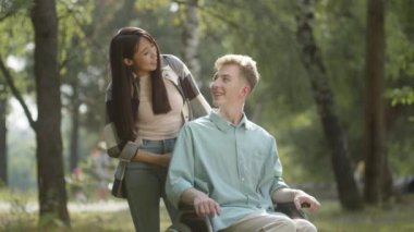 Uzun siyah saçlı sevimli bir Asyalı kadın ve mavi gömlekli engelli genç adam halk parkında kameraya bakıyor ve gülümsüyor. Yüksek kalite 4k görüntü